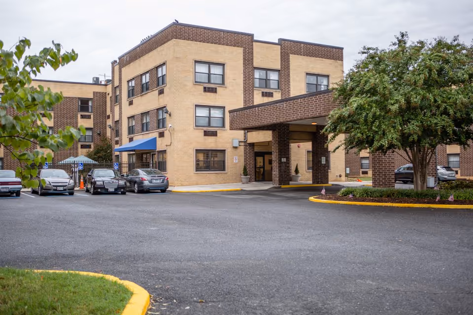 Front entrance and parking area of a multi-story brick rehab and healthcare building with a covered drop-off and parked cars.