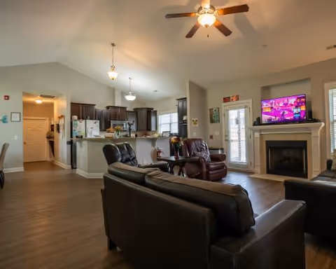 Open communal living room with leather sofas, fireplace, TV, and kitchen in background.