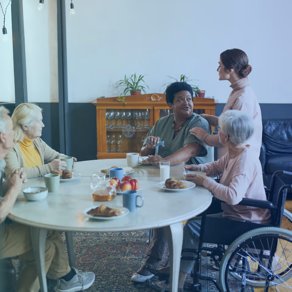 Senior residents dine together at a round table, while a caregiver chats nearby.