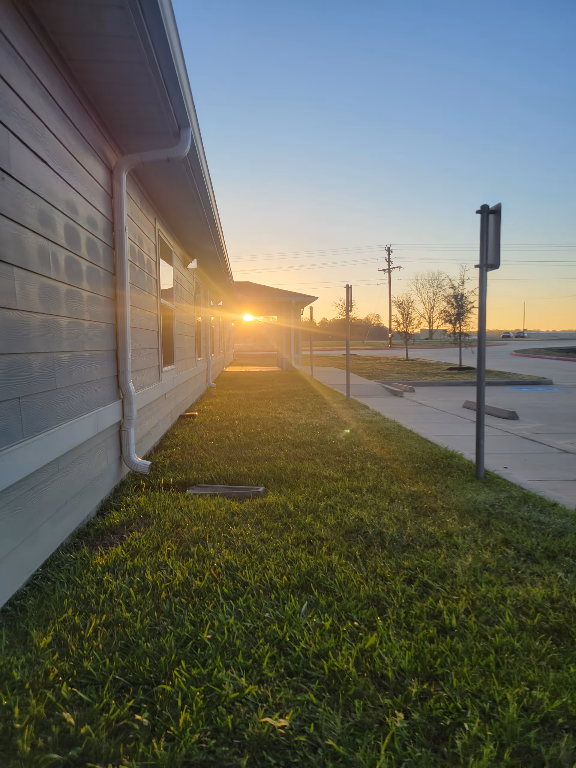 Sunset view along the side of a hospice facility with grass, windows, and sidewalk.