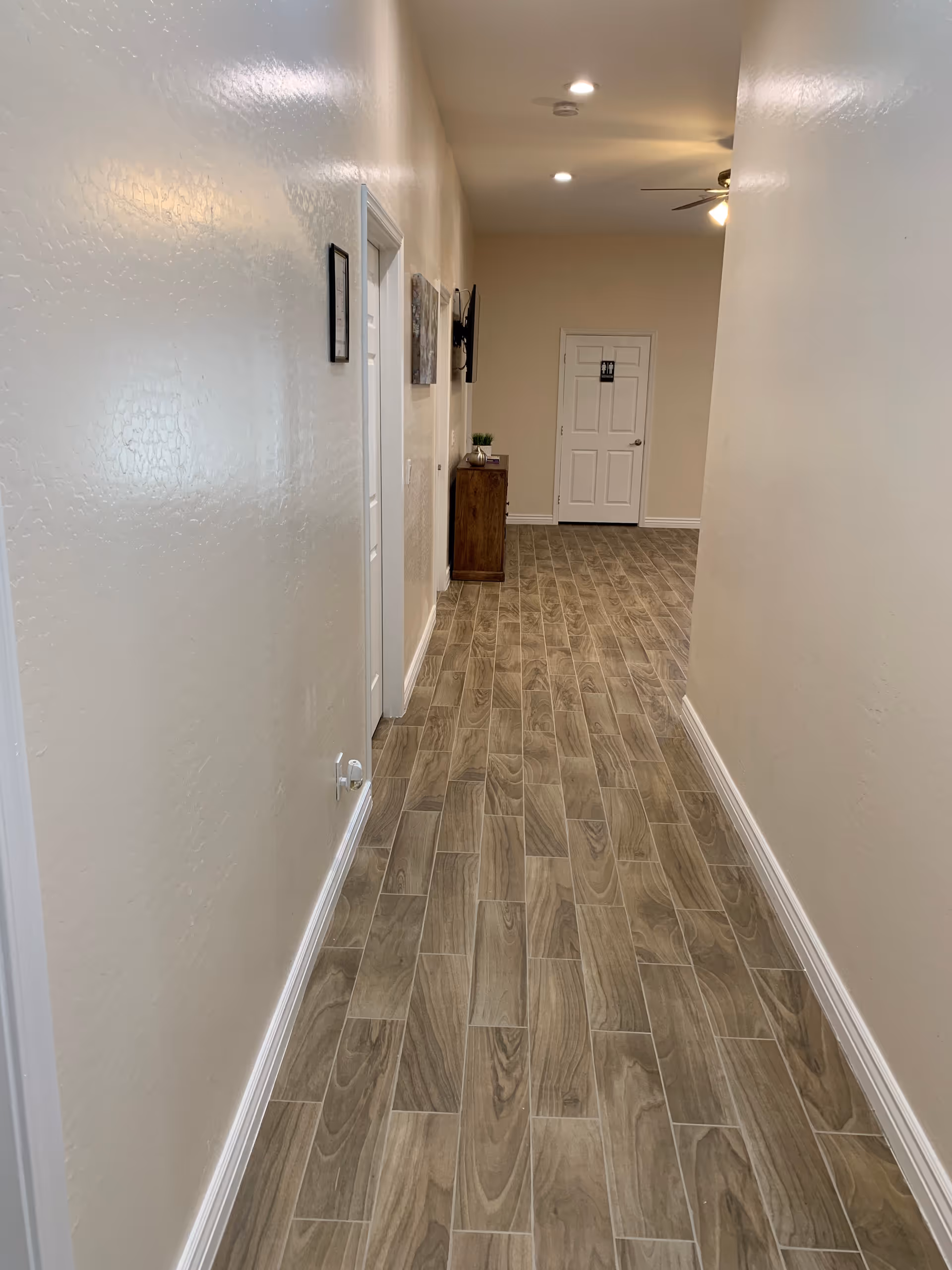 Long interior hallway with wood-look tile floor, beige walls, doors along the left side and a small cabinet at the far end.