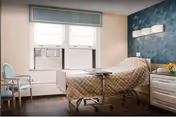 Hospital-style patient room featuring adjustable bed, two chairs, teal accent wall.