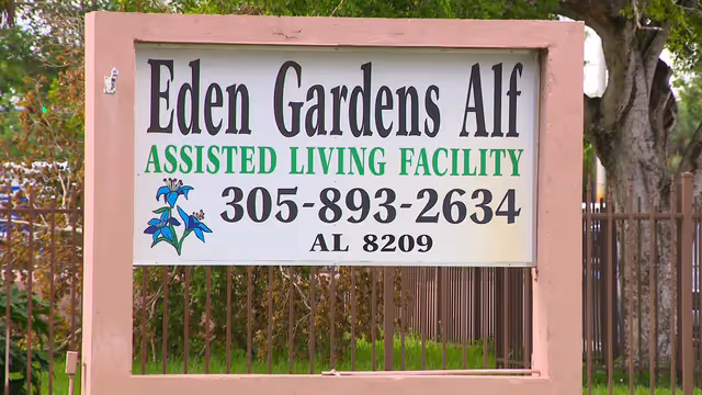 Outdoor sign for 'Eden Gardens ALF Assisted Living Facility' with phone number mounted in a pink frame in front of a fence and trees.