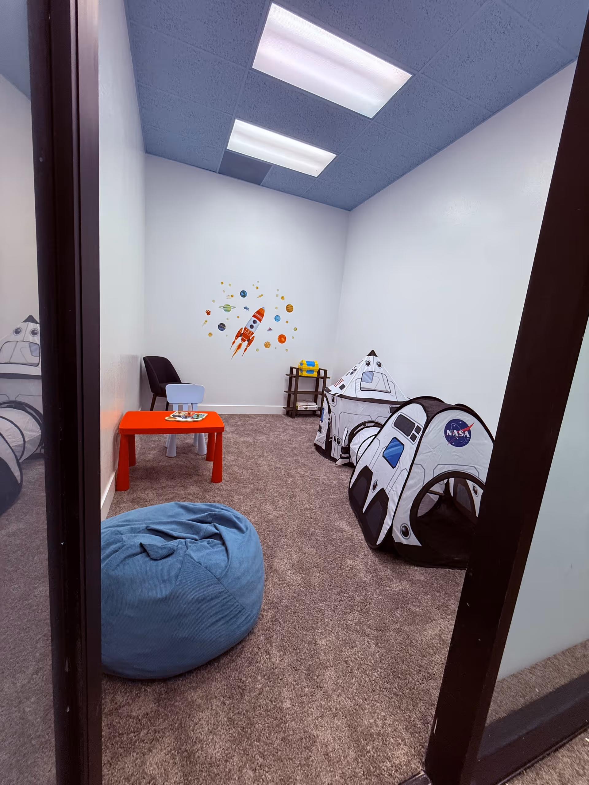 Small playroom with space-themed decals, a red table, chairs, rocket tents, and a bean bag.