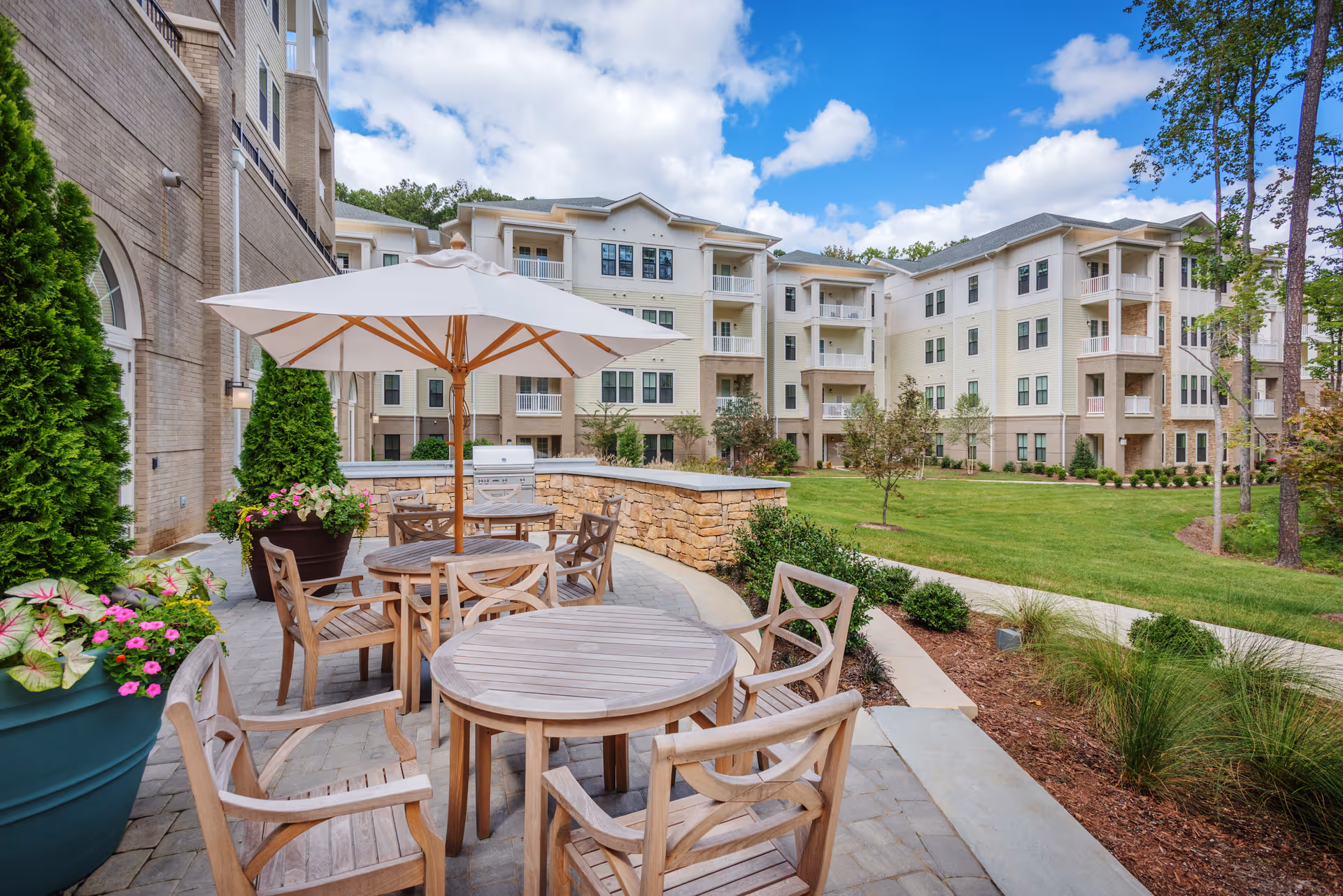 Outdoor patio area at The Templeton of Cary featuring wooden tables and chairs with a large white umbrella, surrounded by potted plants and greenery. In the background, there is a multi-story residential building with balconies and a well-maintained lawn under a partly cloudy sky.