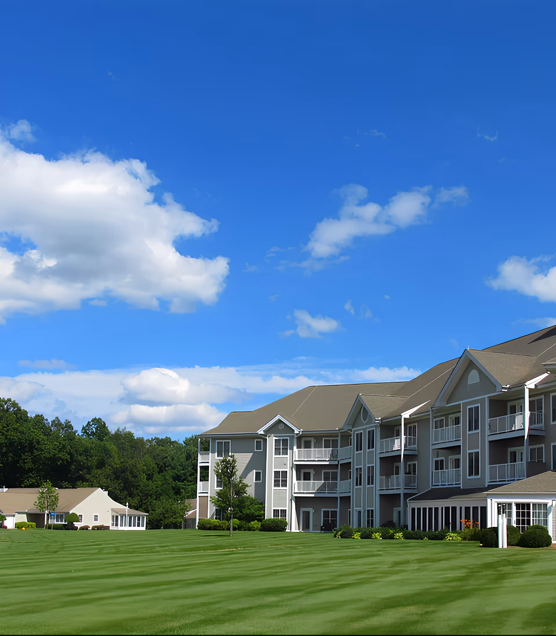 Exterior view of a large senior living facility building with multiple balconies and a well-maintained green lawn under a bright blue sky with scattered clouds.