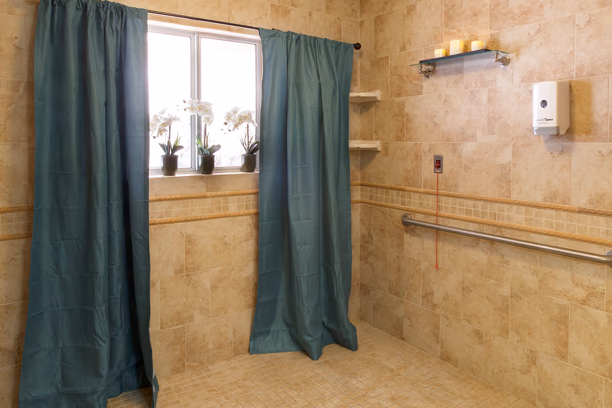 Tile-walled accessible shower area with teal privacy curtains, a window with potted orchids, a grab bar and an emergency pull cord.