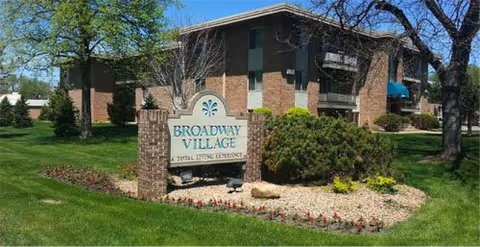 Brick senior living building exterior with a Broadway Village sign amid landscaped lawn.