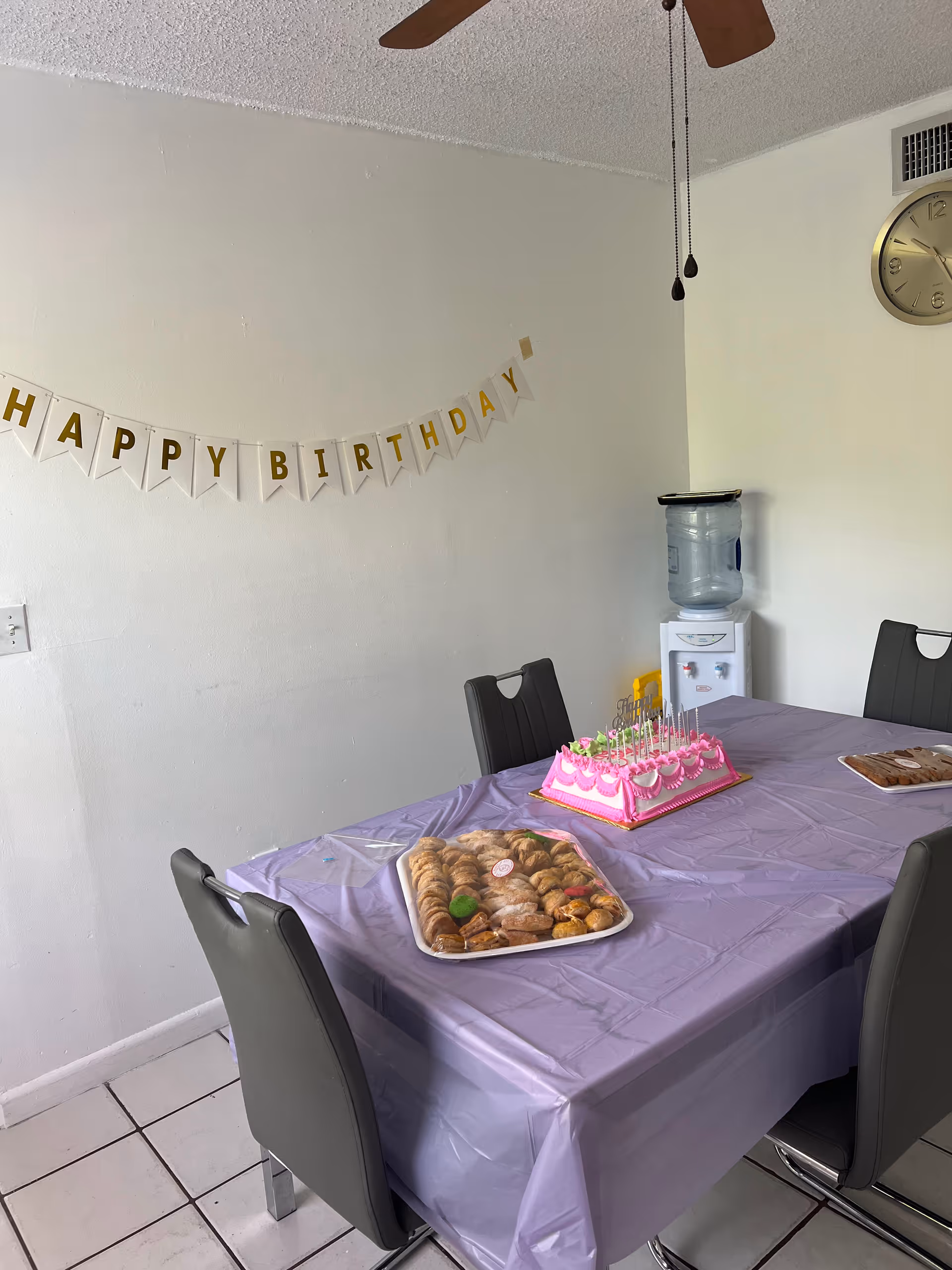 A dining room with a table covered in a purple tablecloth, set with a pink birthday cake with candles and a tray of assorted pastries. There are four gray chairs around the table, a water dispenser in the corner, a wall clock, and a 'HAPPY BIRTHDAY' banner hanging on the wall.