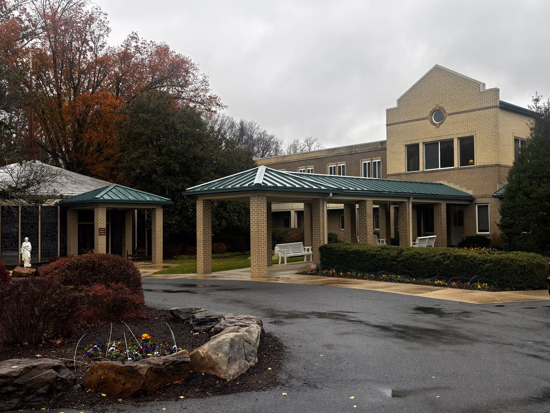 Exterior view of a senior living facility building with a covered entrance supported by brick columns. There are white benches under the covered area and landscaped bushes and flowers around the driveway. Trees with autumn foliage are visible in the background under a cloudy sky.