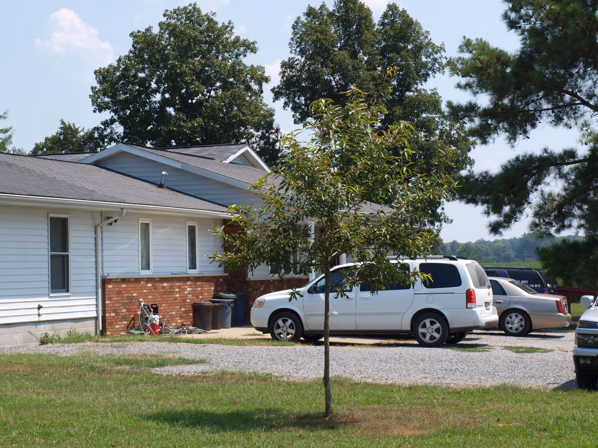 Exterior view of a single-story building with white siding and a brick lower facade, a small tree in the foreground, several parked cars on a gravel driveway, and large trees in the background under a partly cloudy sky.