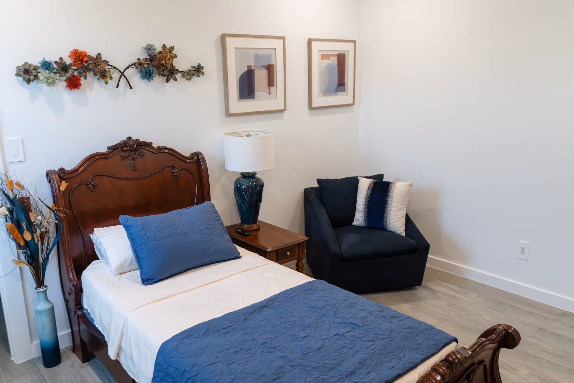 A cozy bedroom features a carved wooden bed, blue bedding, nightstand lamp, and chair.