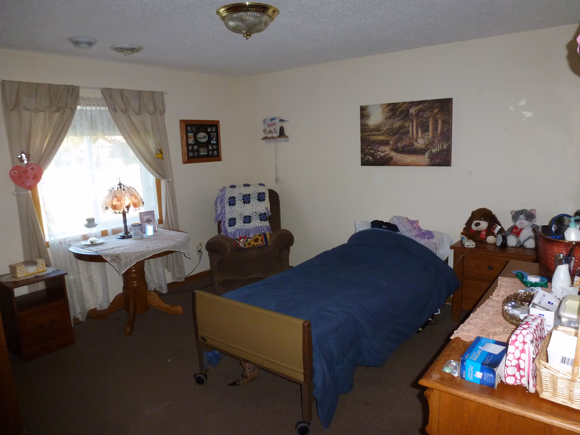 Cozy resident bedroom with a bed, dresser, table, and window curtains in an assisted living facility.