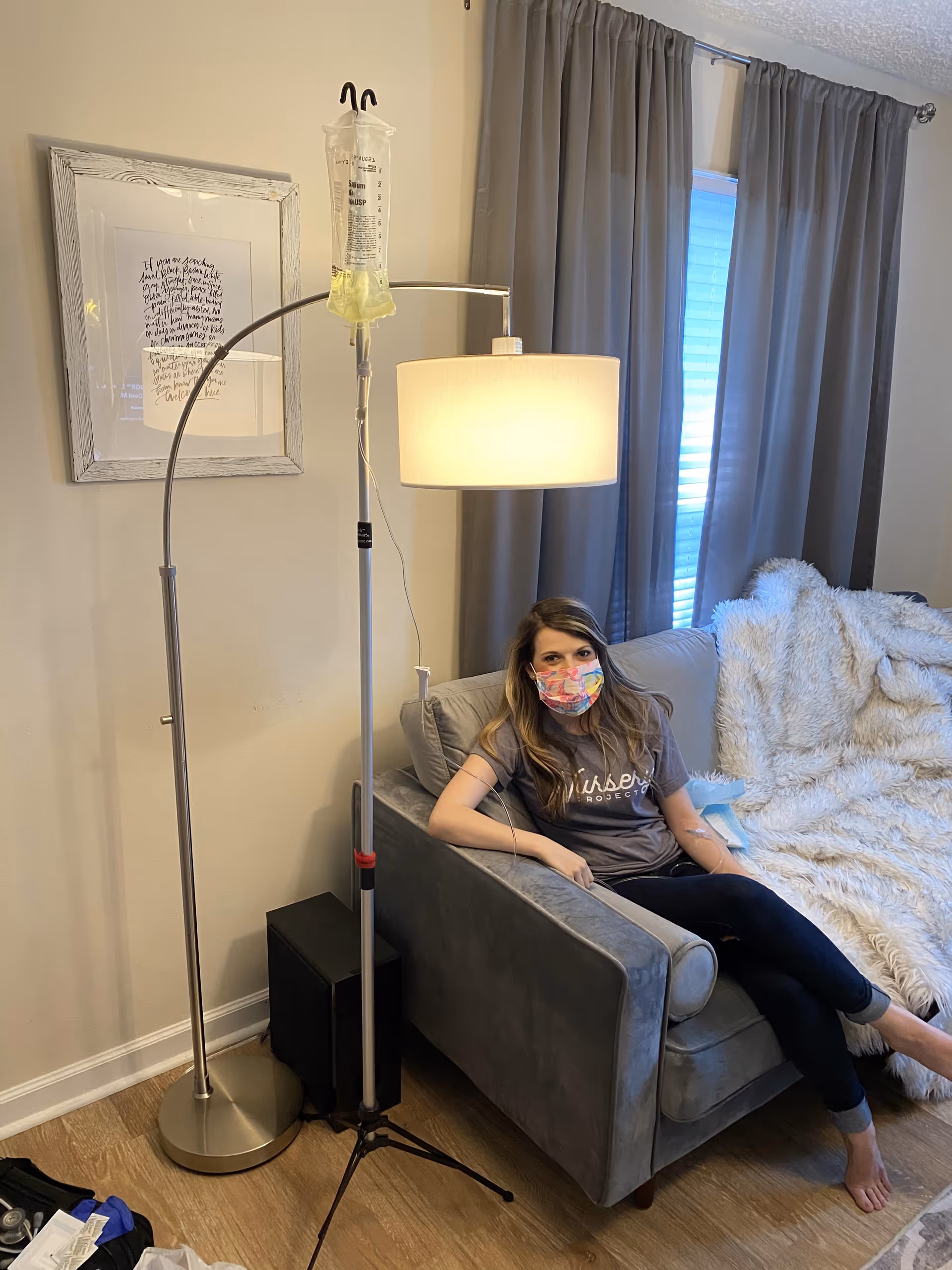 Masked resident sits on a gray sofa in a living room with IV pole and lamp.