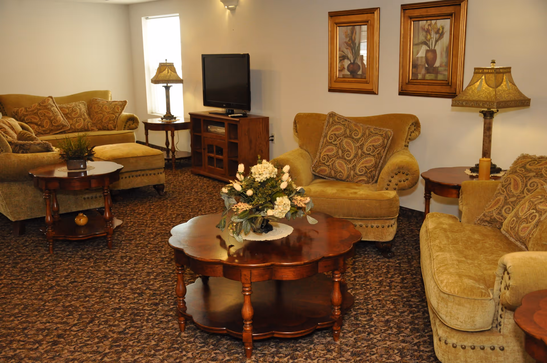 A cozy living room with beige upholstered sofas and armchairs featuring patterned cushions. There are wooden side tables with decorative lamps and a wooden coffee table with a floral arrangement in the center. A flat-screen TV is placed on a wooden TV stand against the wall, which is decorated with two framed floral paintings.
