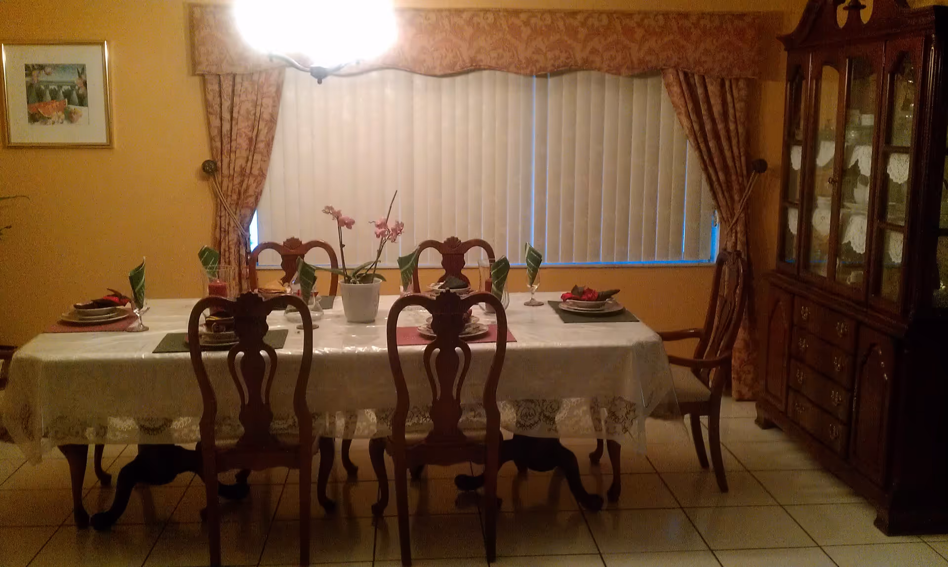 Formal dining room with a long set table, wooden chairs, a china cabinet, and a window with vertical blinds.