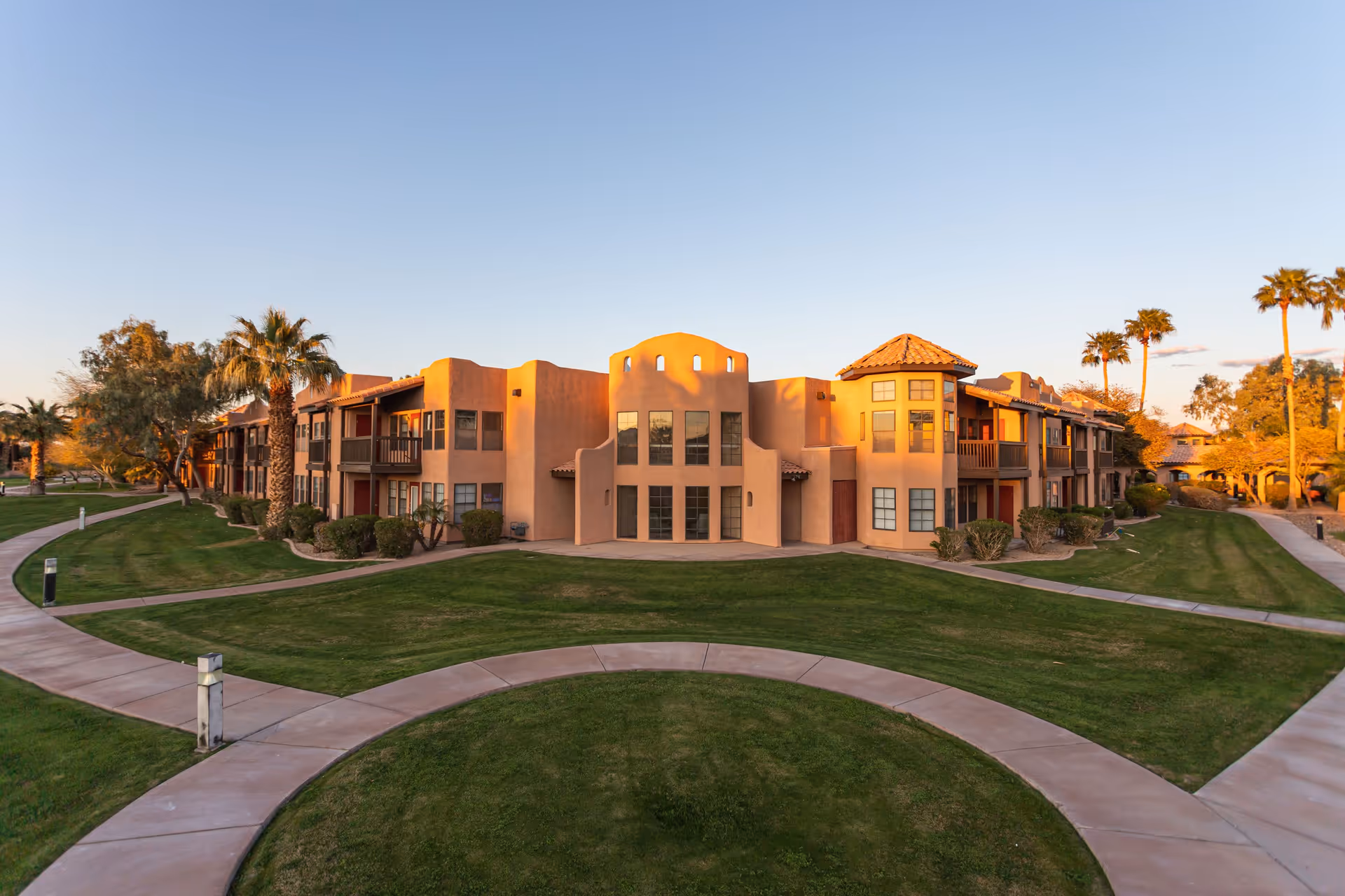 Exterior view of a senior living facility building with southwestern architectural style, surrounded by well-maintained green lawns, palm trees, and curved walkways under a clear sky at sunset.