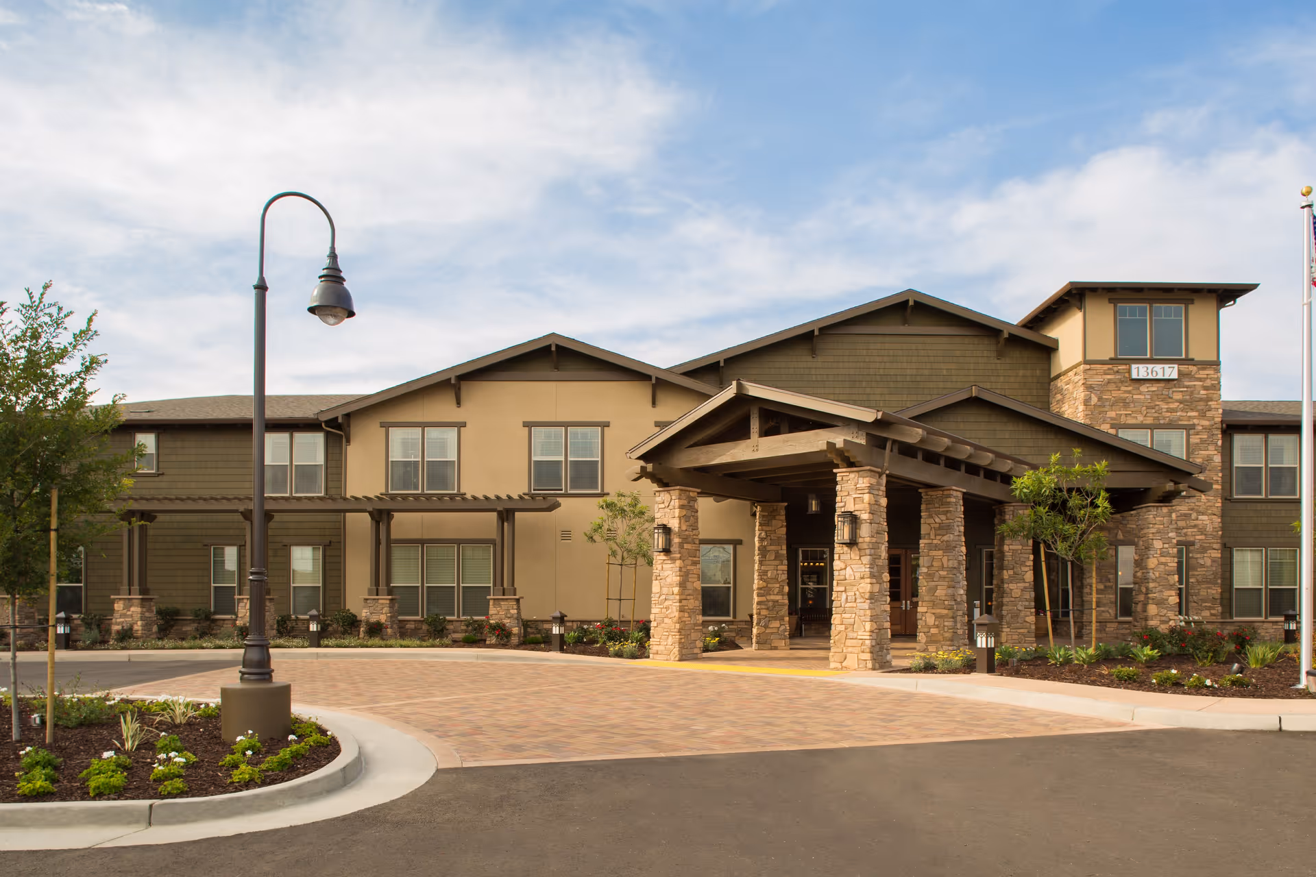 Front exterior view of Oakmont of Whittier senior living facility with a covered entrance supported by stone pillars, landscaped garden beds, a street lamp, and a clear sky in the background.