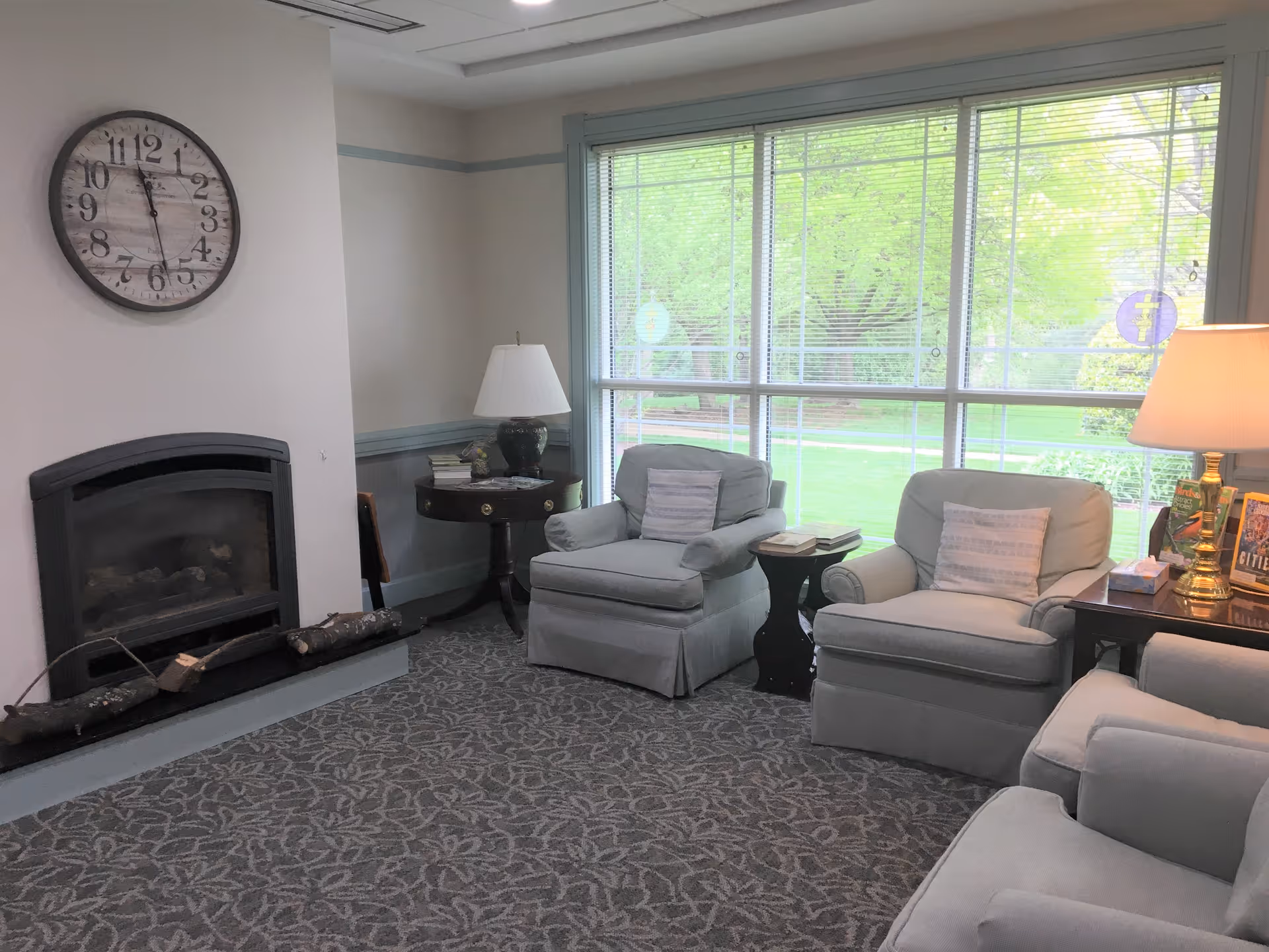 Bright seating area with cushioned armchairs and side tables in front of a large window and a fireplace.