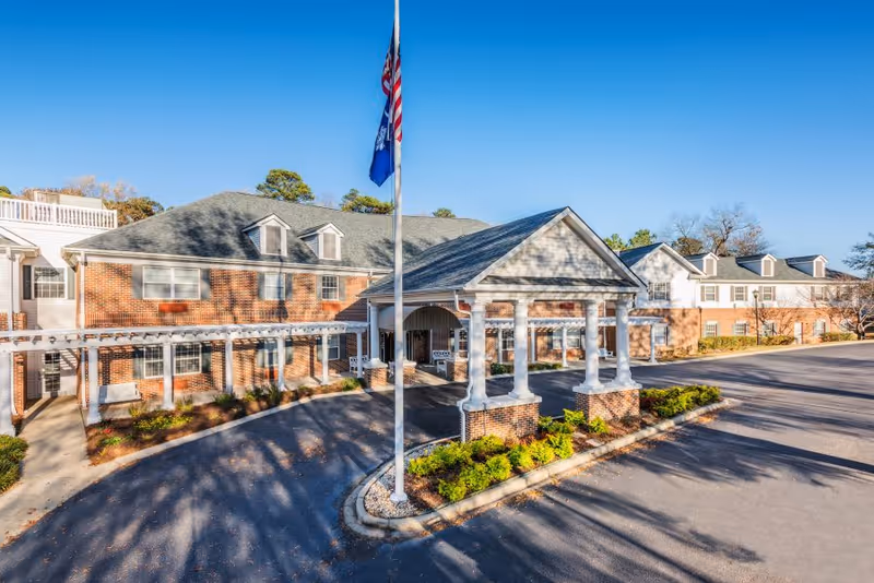 Exterior view of Oakleaf Village of Lexington, showing a large brick building with white columns at the entrance, a covered driveway, landscaped bushes, and two flags on a flagpole under a clear blue sky.