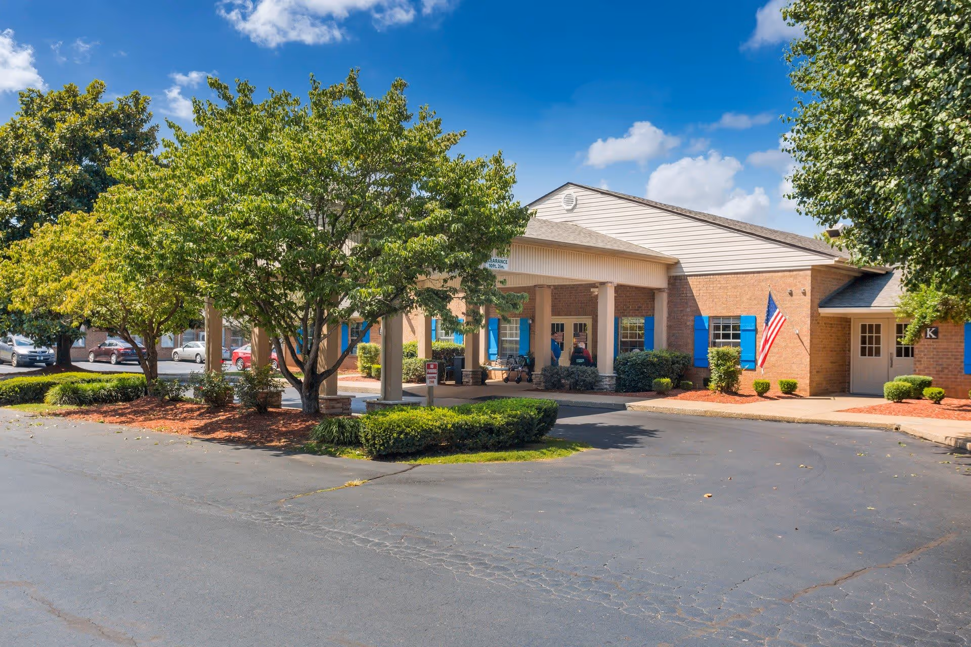 Exterior view of Charter Senior Living of Bowling Green showing the entrance with a covered drop-off area, surrounded by trees and landscaping under a blue sky with some clouds. Several parked cars are visible in the background.