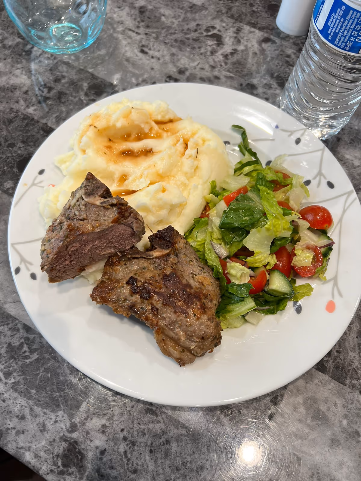 Plate with mashed potatoes, two pieces of cooked meat, and a side salad on a marble table with a water bottle and glass.