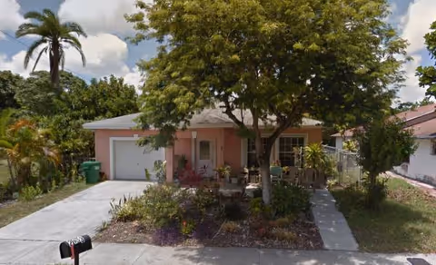 A single-story pink house with a driveway, front yard, and a large tree.