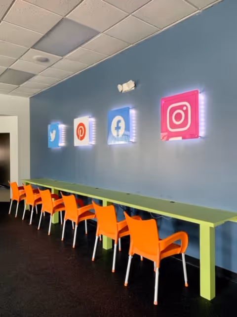Long lime-green table with orange chairs along a blue wall featuring illuminated social media icons.