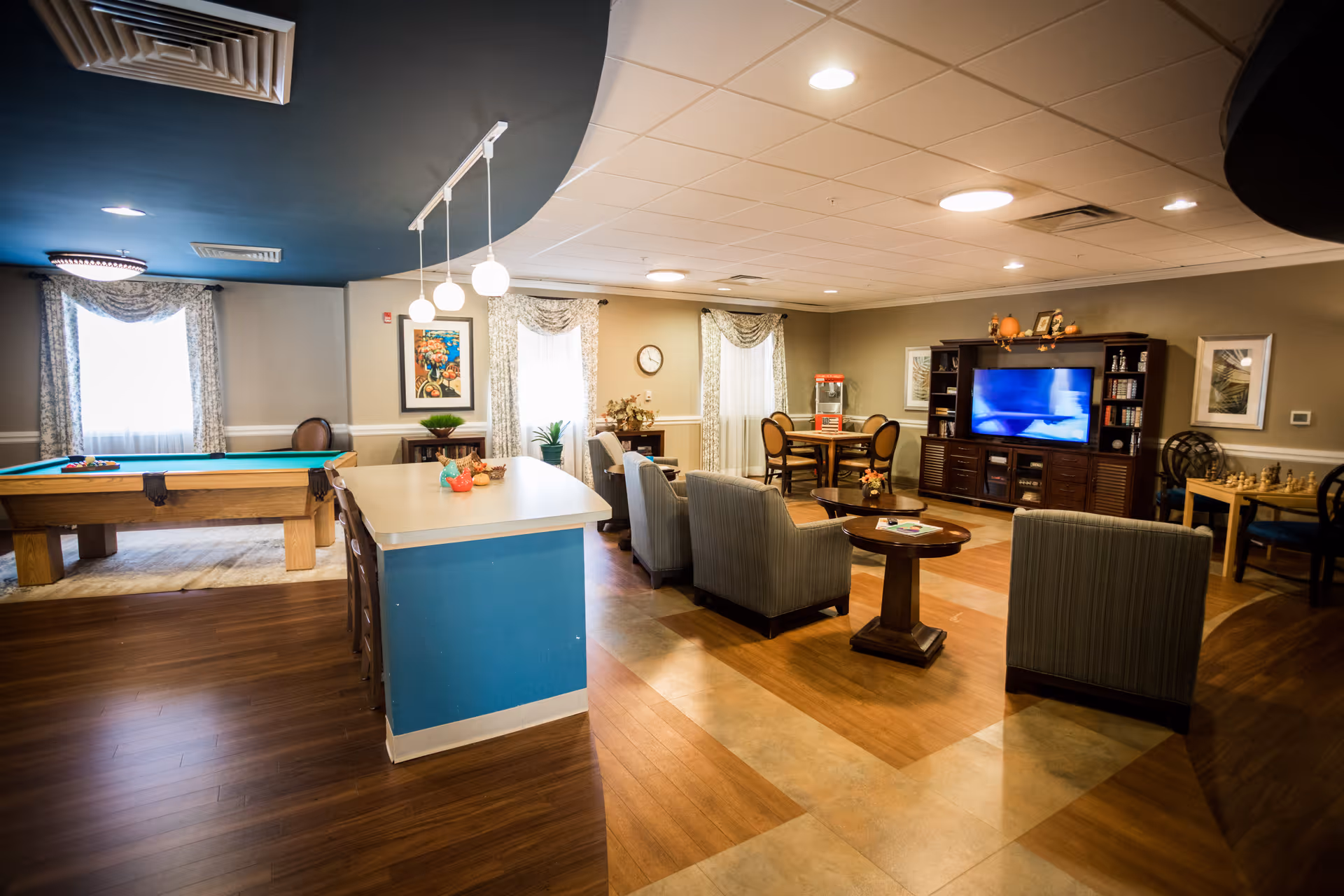 A spacious and well-lit common area in a senior living facility featuring a pool table, a kitchen island with chairs, several armchairs arranged around a coffee table, a large TV on a wooden entertainment center, and a small dining table with chairs. The room has wood and tile flooring, large windows with curtains, and decorative artwork on the walls.