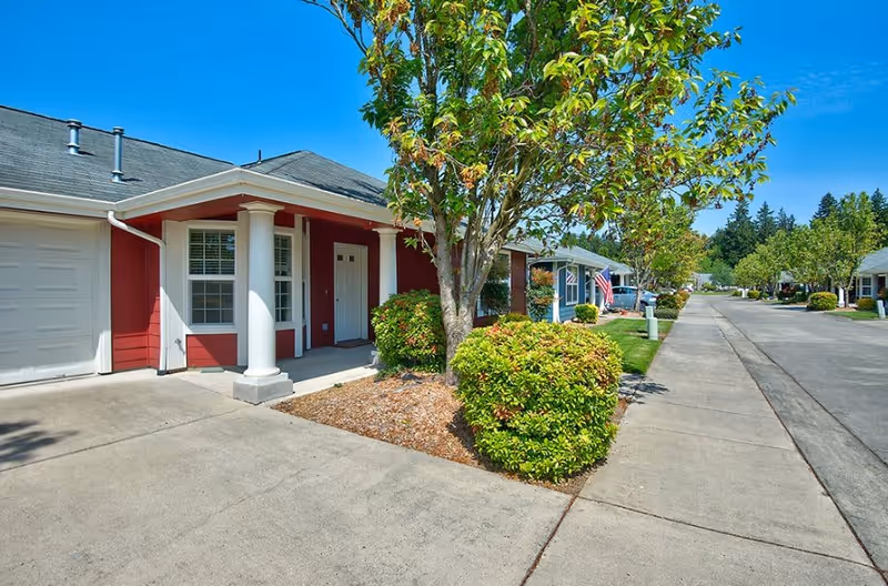 Exterior view of a residential unit at Parkland Village Retirement Community featuring a red house with white columns, a garage, a tree, and neatly trimmed bushes along a sidewalk on a sunny day.