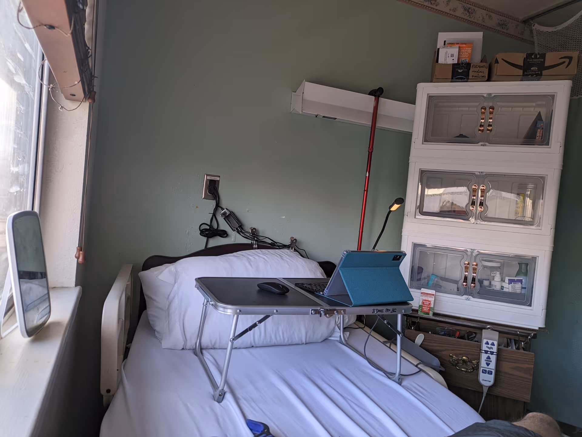 Small hospital-style bedroom with a bed, laptop tray, cabinet, and medical supplies