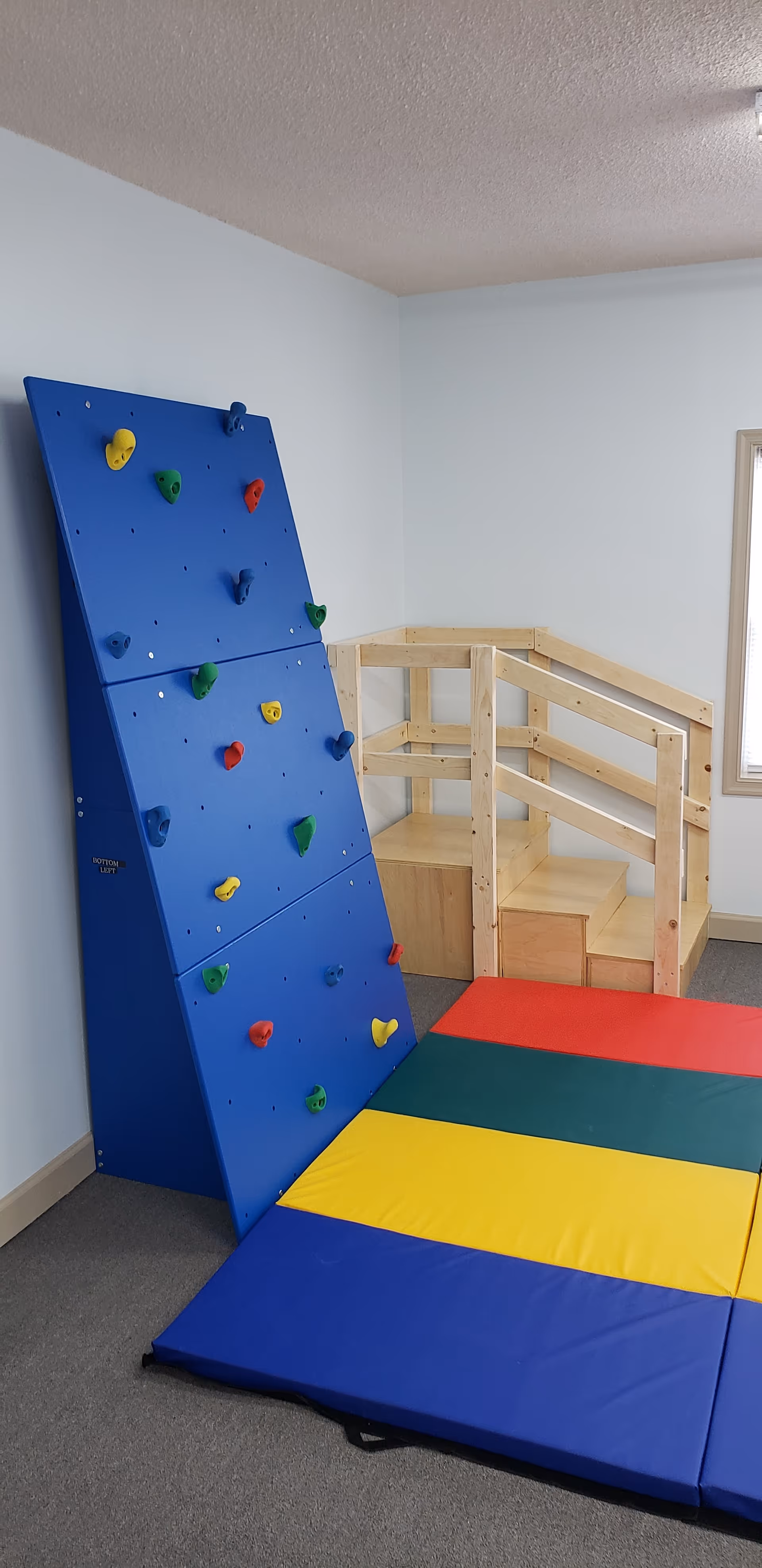 Blue climbing wall with colorful holds, padded mats, and a wooden play structure.