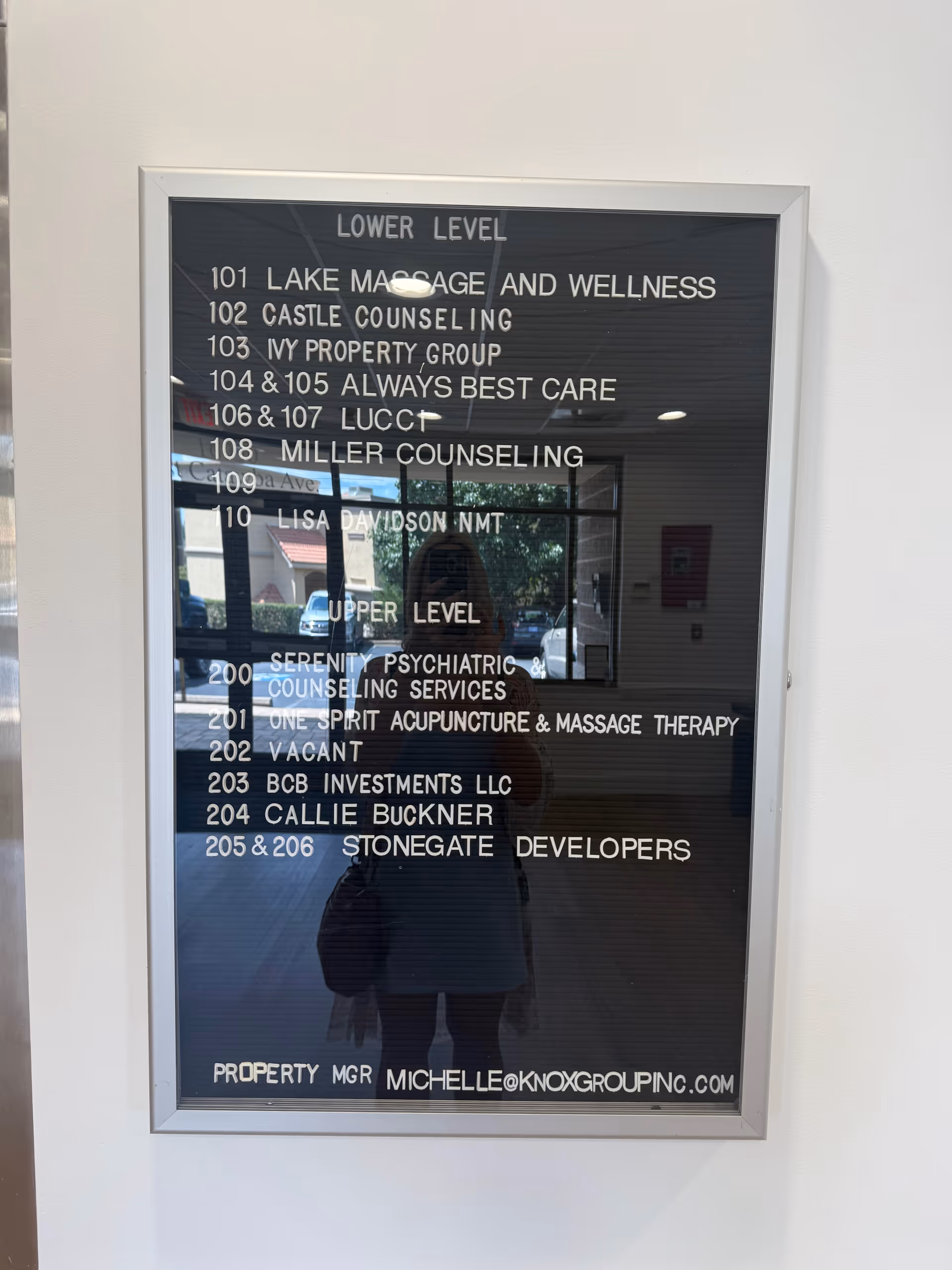 Interior wall directory plaque listing offices and suites on the lower level, reflection visible.