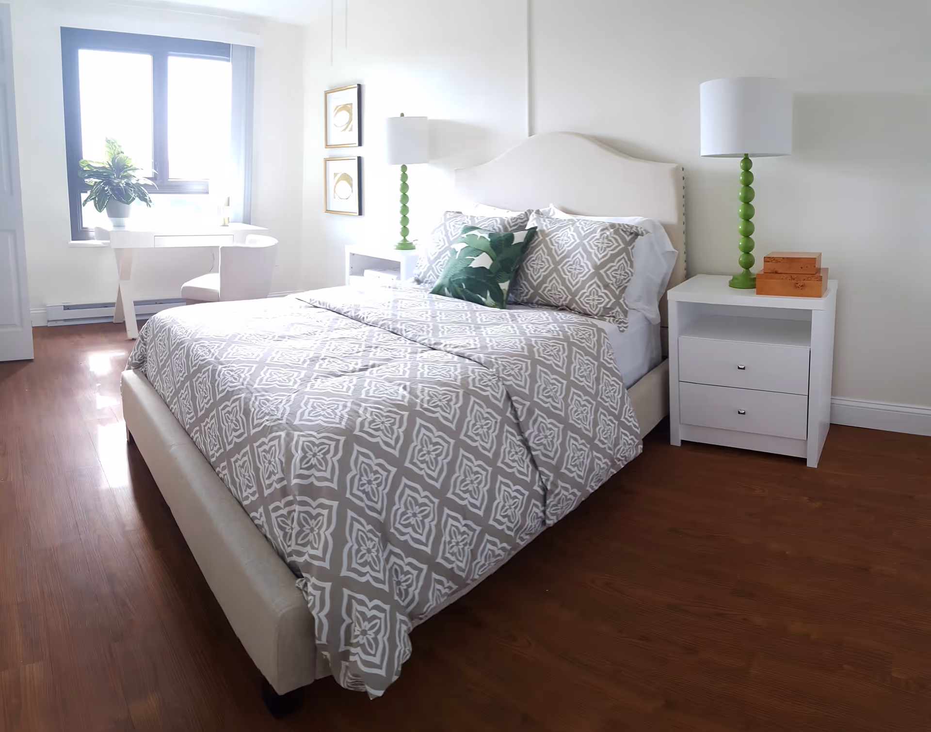 A bright bedroom with a large window letting in natural light. The room features a bed with a beige upholstered headboard, patterned gray and white bedding, and a green decorative pillow. On either side of the bed are white nightstands, each with a green lamp. There is a white desk and chair near the window, and two framed artworks hang on the wall beside the bed. The floor is wooden.