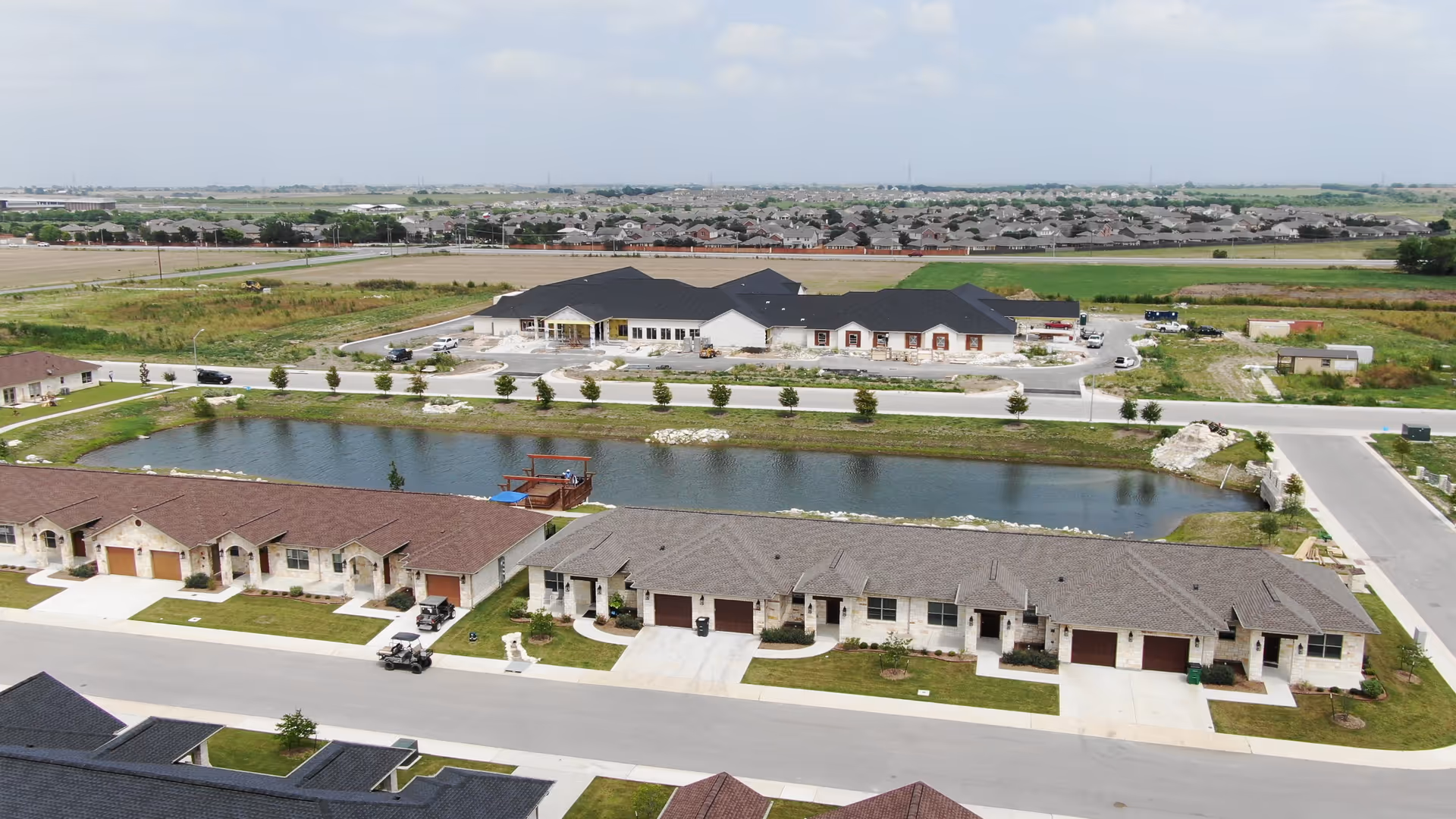 Aerial view of a senior living community with single-story row homes bordering a pond and a larger community building across the water.