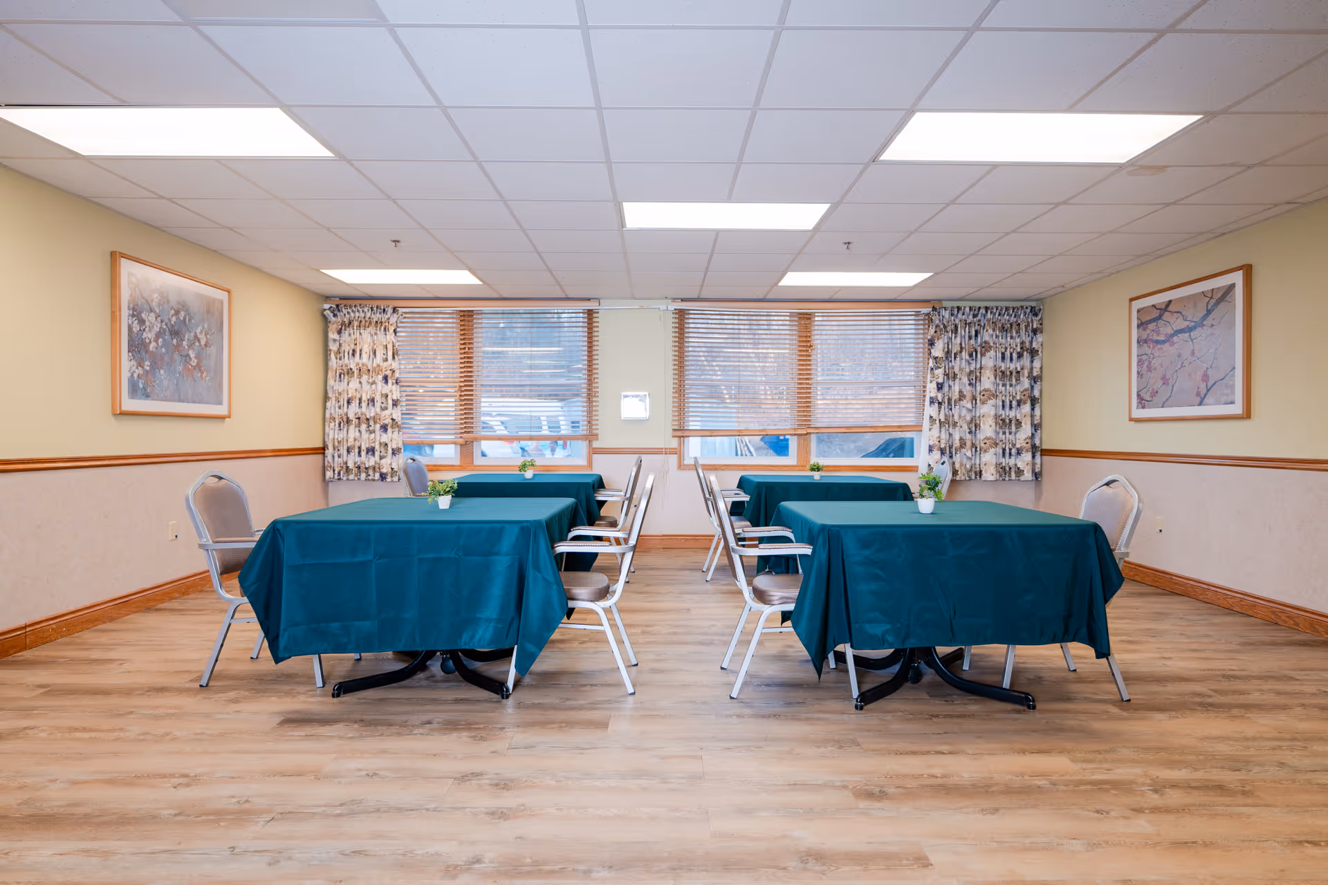 Senior living dining room with teal tablecloths, metal chairs, and windows.