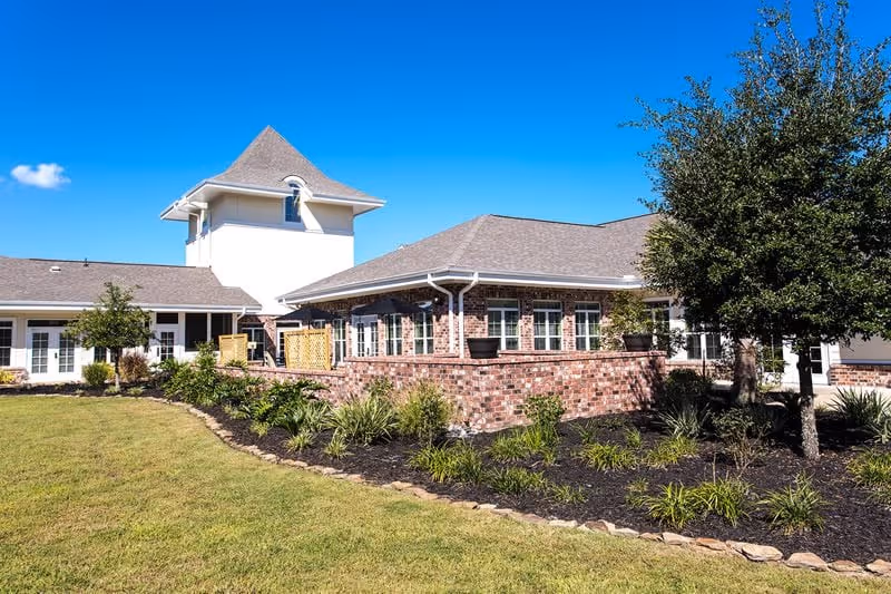 Brick-faced senior living facility building with a landscaped lawn, trees, and a bright blue sky.