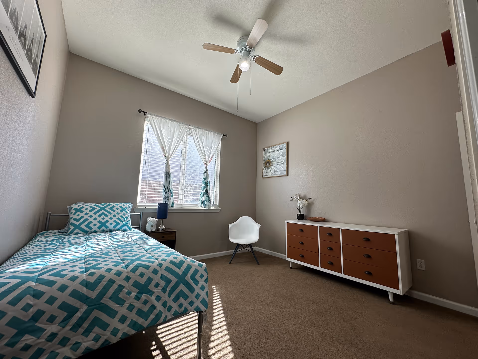 Small bedroom with single bed, dresser, chair, and window with white curtains under a ceiling fan.