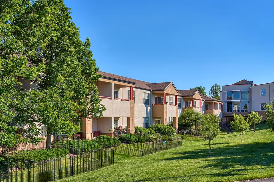 Sunlit exterior view of a senior living campus with beige buildings, balconies, and green lawns.