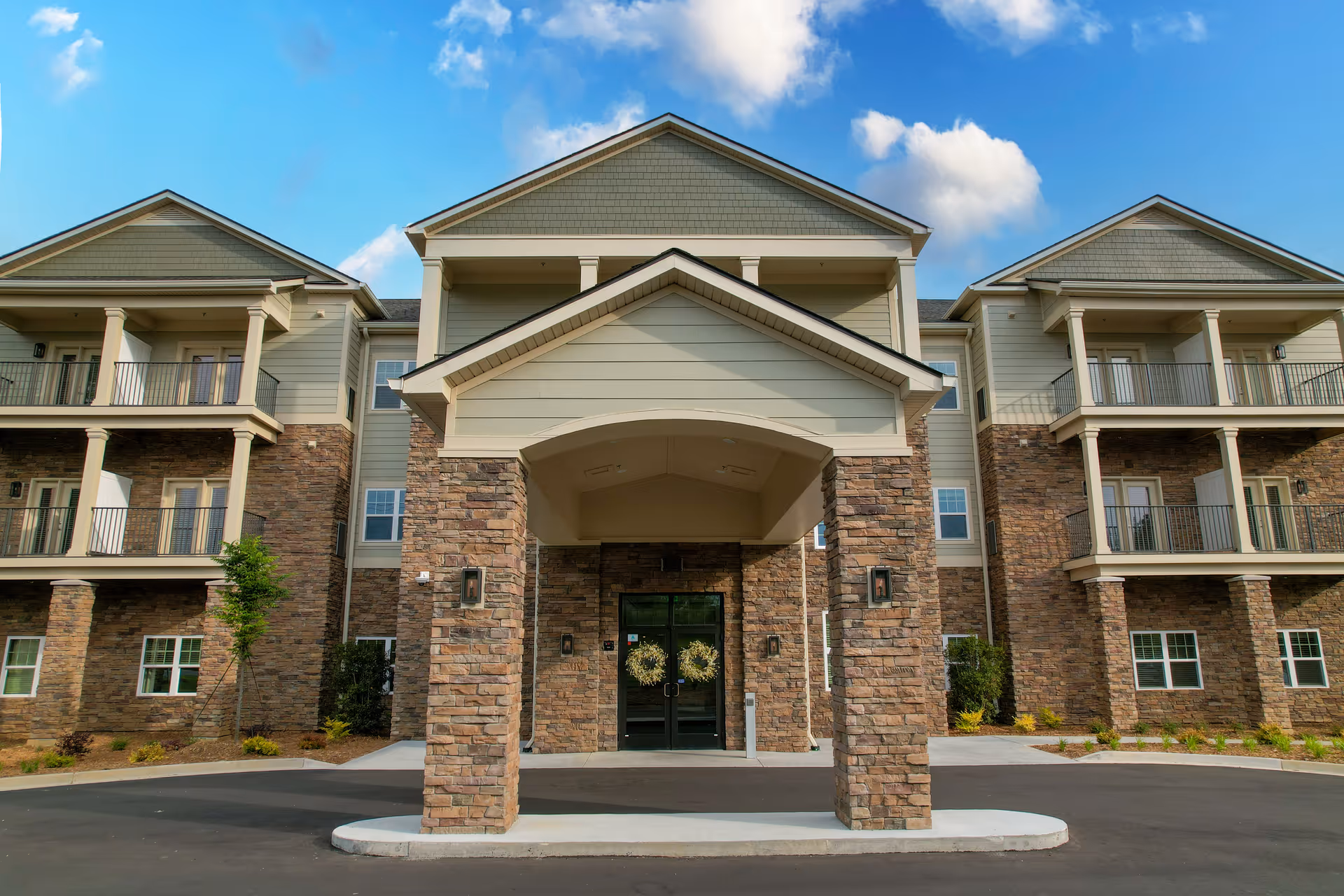 Front exterior view of a three-story senior living facility building with stone and beige siding, balconies, and a covered entrance with stone pillars under a blue sky with some clouds.