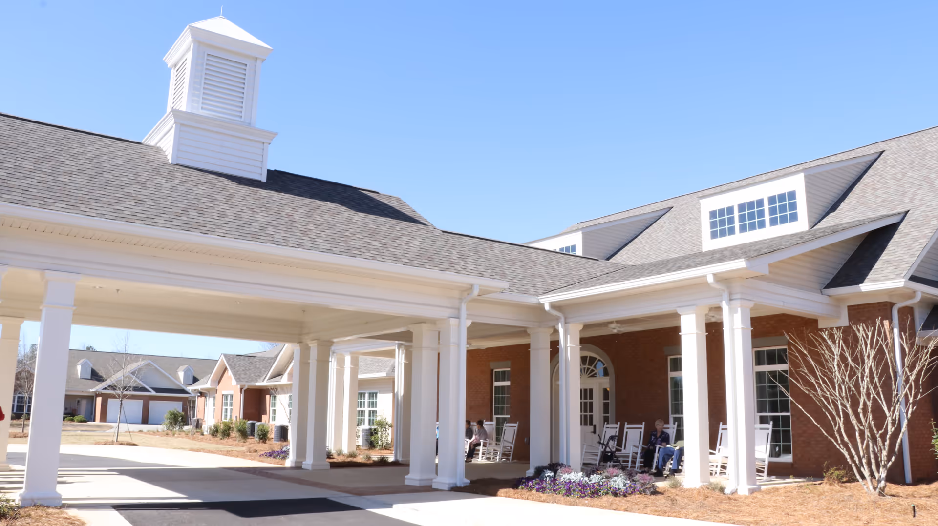 Exterior view of The Cottages of Milledgeville senior living facility entrance with a covered drop-off area supported by white columns, brick walls, and a few people sitting on rocking chairs on the porch under a clear blue sky.