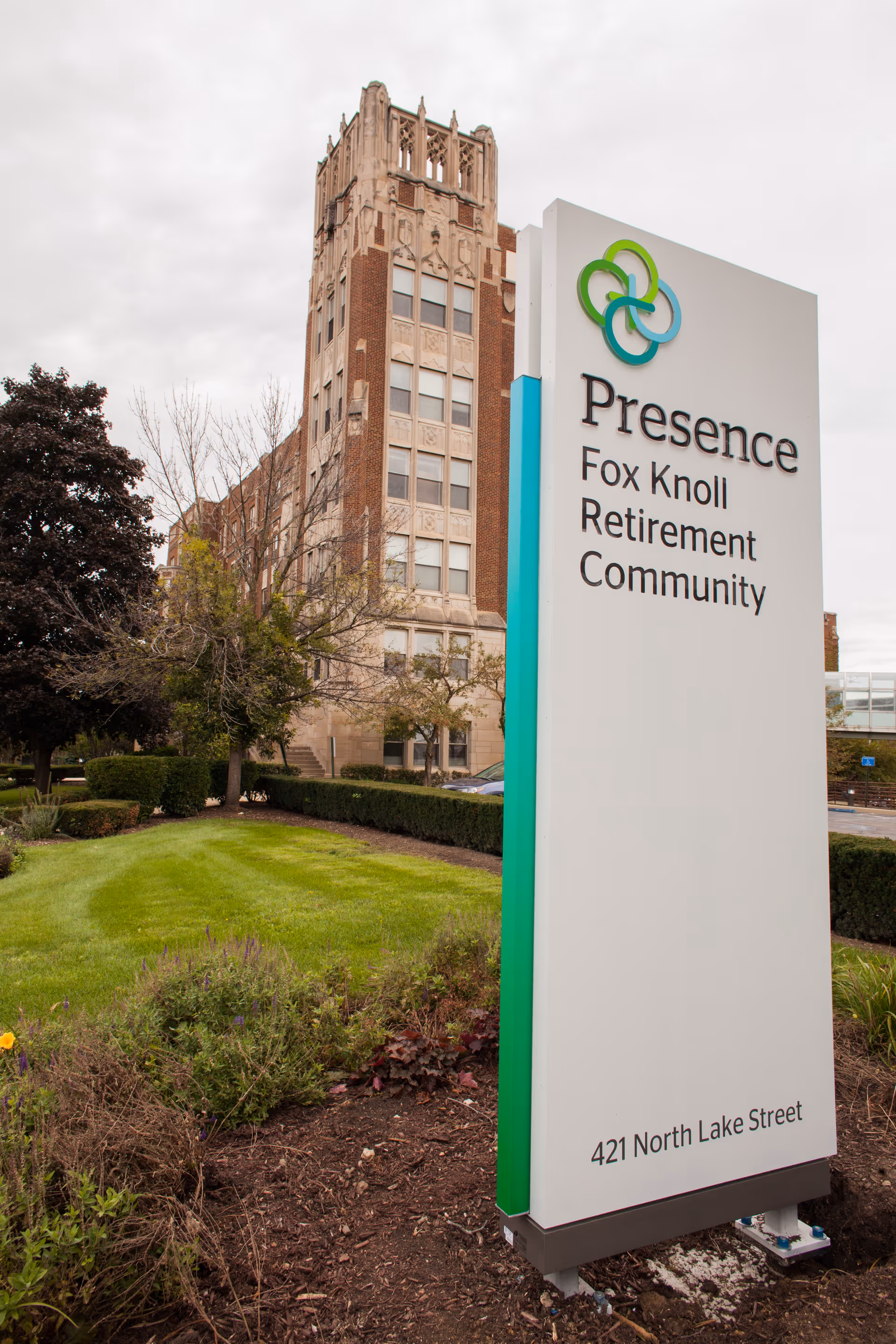 Tall exterior sign for Presence Fox Knoll Retirement Community with brick building behind