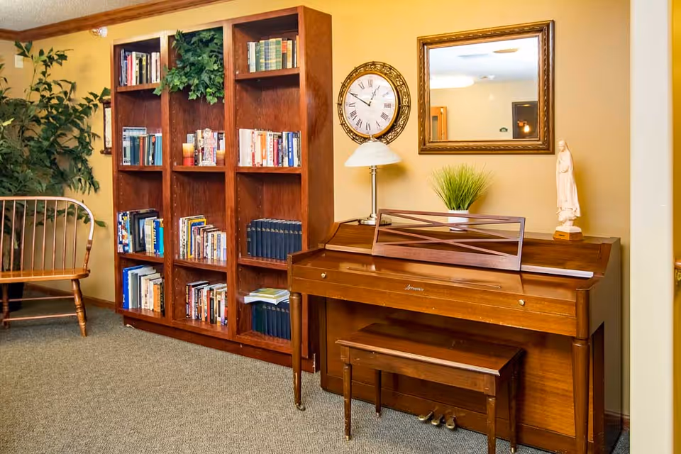 A cozy interior room with a wooden upright piano and matching bench. Above the piano is a round wall clock and a rectangular mirror. On top of the piano are a small lamp, a potted plant, and a statue. To the left of the piano is a tall wooden bookshelf filled with books and some decorative greenery. A wooden bench with a curved backrest is positioned against the wall near the bookshelf. The walls are painted a warm beige color and the floor is carpeted.