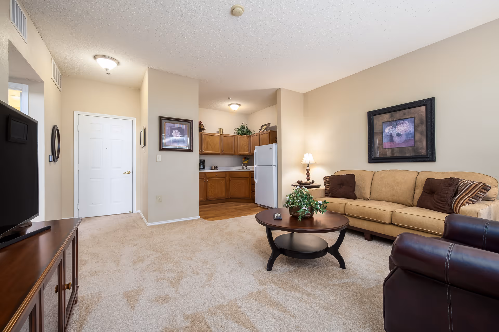 Cozy senior living room with beige sofa, coffee table, TV, and kitchenette.