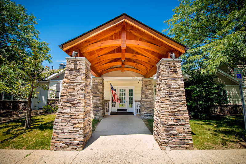 Sunny exterior entrance with stone columns and a wooden canopy at a senior living facility.