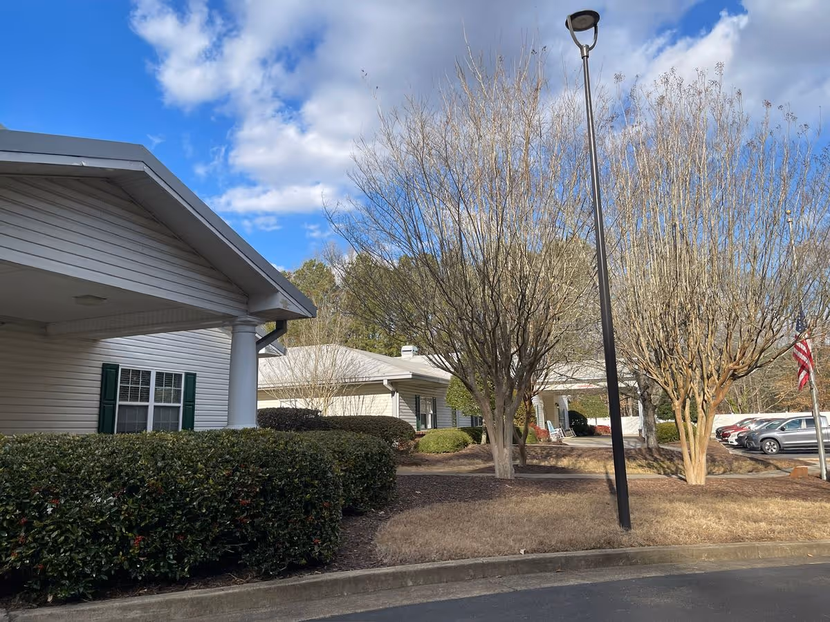 Exterior view of a single-story senior living building with trimmed hedges, leafless trees, a lamppost, and parked cars under a blue sky.