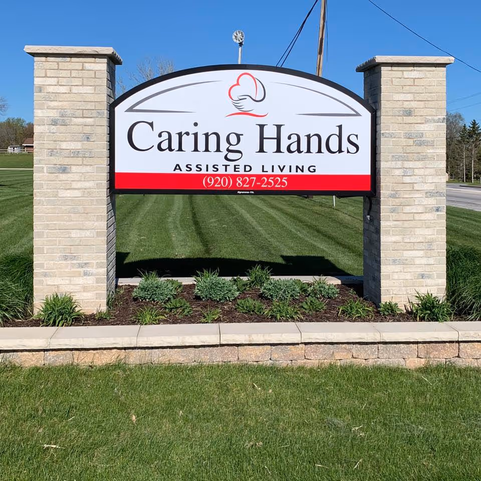 A large outdoor sign for Caring Hands Assisted Living mounted between two beige brick pillars with a landscaped area of green plants and grass in front, under a clear blue sky.
