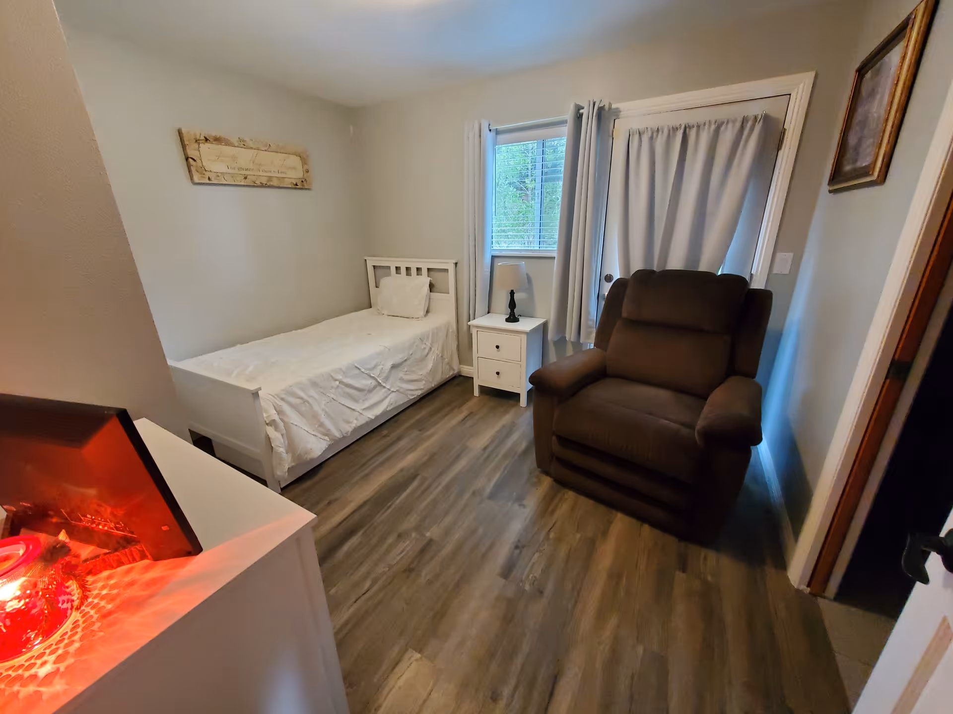 A small bedroom with a single white bed against the left wall, a white nightstand with a lamp next to a window with white curtains, and a large brown recliner chair near a door with a curtain. The floor is wood, and there is a framed picture on the right wall and a decorative sign on the left wall.