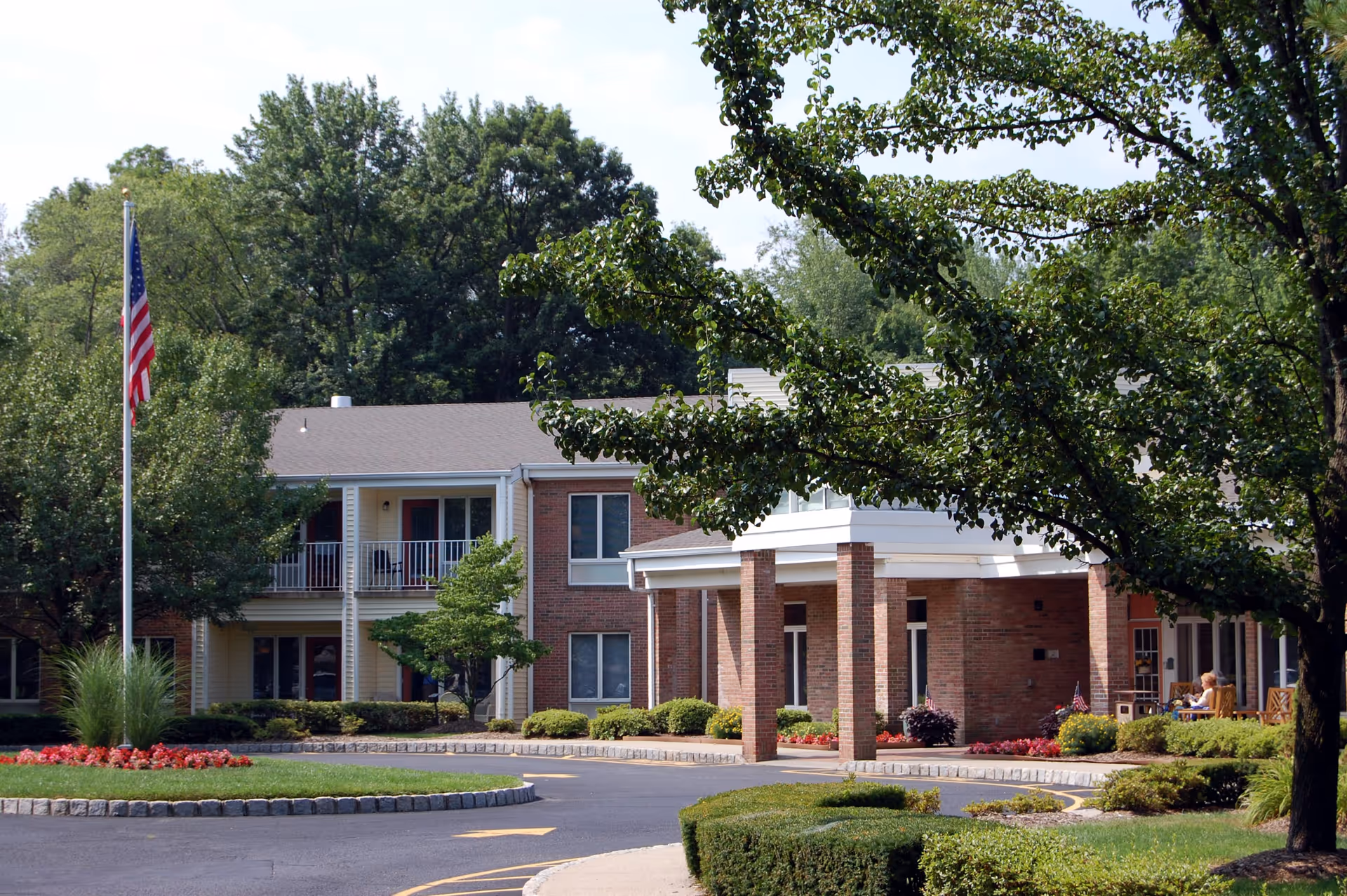 Exterior view of a two-story assisted living facility building with brick and light-colored siding, surrounded by well-maintained landscaping including trees, bushes, and flower beds. An American flag is displayed on a flagpole near the entrance, and a covered driveway leads to the main entrance where a few people are seated on benches.