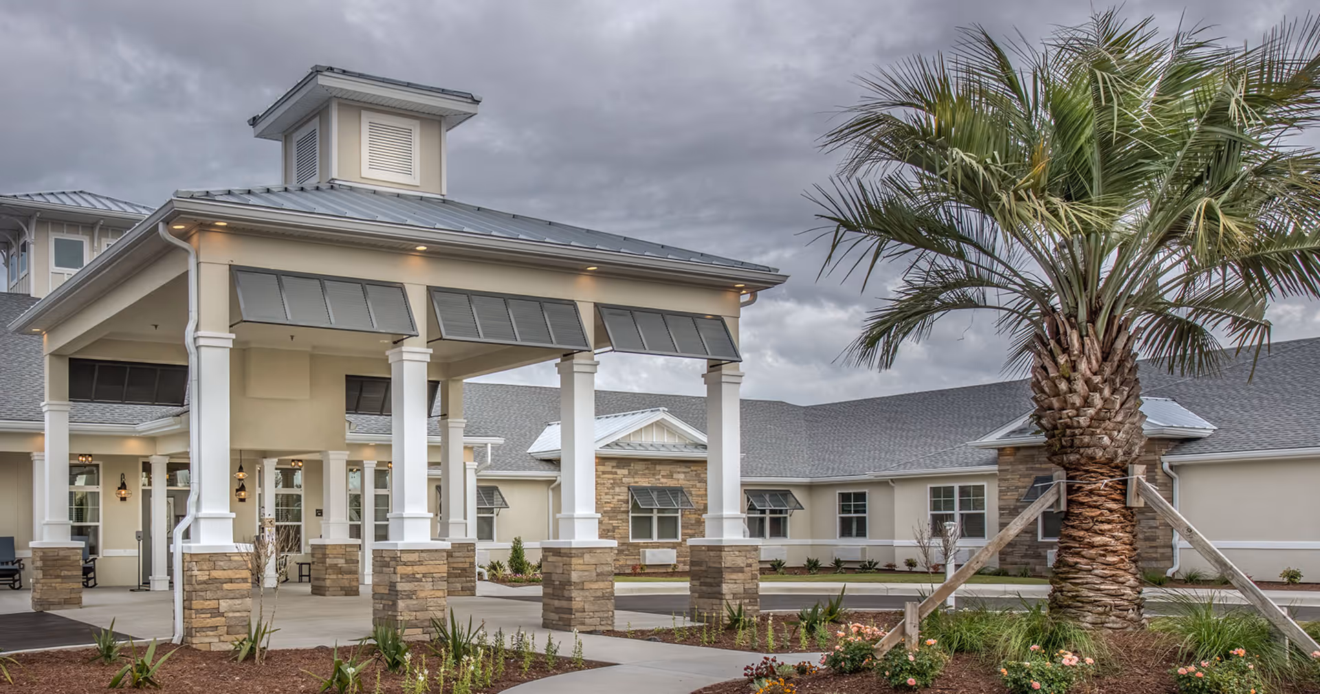 Exterior view of The Addison of Narcoossee senior living facility showing a covered entrance with white columns and stone bases, a large palm tree in the landscaped front area, and a cloudy sky overhead.